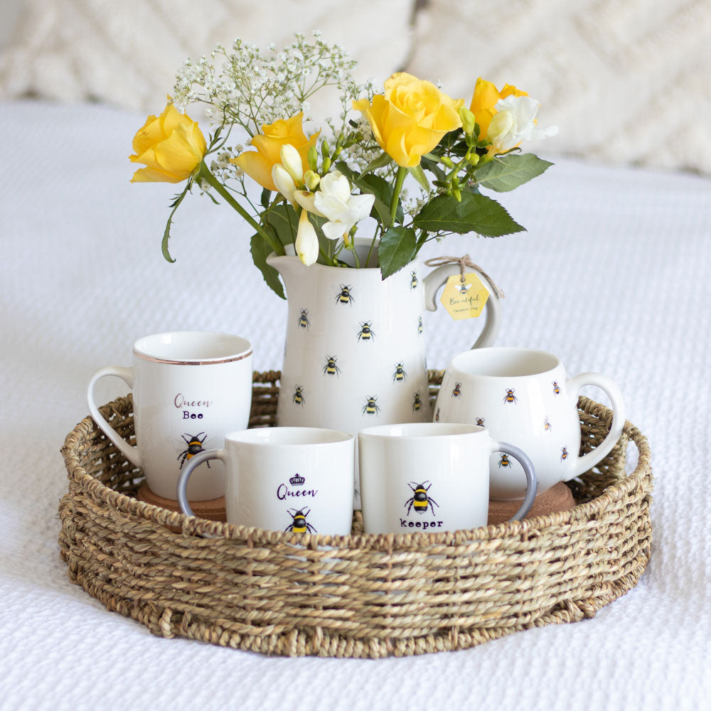 Bee-themed ceramic pitcher and mugs on a woven tray.